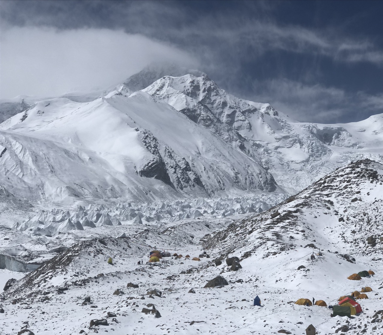Base camp tents spread below a glacier under heavy cloud